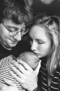 Ein Foto von einem Babyshooting. Es ist ein schwarz/weiß Foto. Die beiden Eltern halten ihr Baby in den Armen, während sie eng beinander stehen. Die Mutter gibt dem Baby ein Küsschen auf das Köpfchen.
