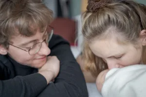 Die Eltern liegen im Bett und haben das Baby vor sich liegen. Die Mutter gibt dem Baby einen Kuss während der Vater ganz beseelt seine Familie anguckt.