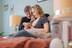 Ein Foto von einem Babyshooting. Es ist ein schwarz/weiß Foto Zu sehen ist eine Familie mit Vater, Mutter und Baby, welches gerade gestillt wird. Die Eltern lehnen die Köpfe aneinander, während die Mutter ihr Baby hält.