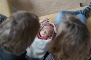Das Foto wurde von oben gemacht. Die Eltern sitzen nebeneinander, halten das Baby im Schoß und streicheln es über den Kopf.