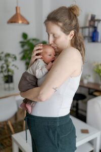 Es handelt sich um ein buntes Foto bei einem Babyfotoshooting. Es ist eine Mutter mit ihrem Baby zu sehen. Sie küsst es liebevoll auf den Kopf und stützt liebevoll den Kopf mit ihrer Hand.