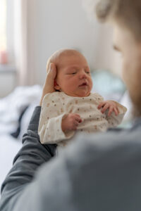 Es handelt sich um ein buntes Foto bei einem Babyfotoshooting. Es ist ein Porträt von einem Baby, welches gerade von seinem Vater getragen wird. Das Foto ist so geschossen, dass im Vordergrund noch verschwommene Teile des Vater zu sehen ist und Schulter und Kopf das Bild Rahmen. Mittig ist das Baby zu sehen.