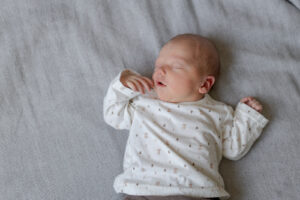 Es handelt sich um ein buntes Foto bei einem Babyfotoshooting. Es ist ein Porträt von einem Baby, welches gemütlich auf dem Bett liegt und zu schlafen scheint. Dabei liegt ein Ärmchen auf der Seite, während das andere an der Wange ist.